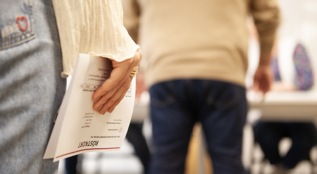 Närbild på en persons hand som håller i ett röstkort. Framför hen står en annan person i kö.
