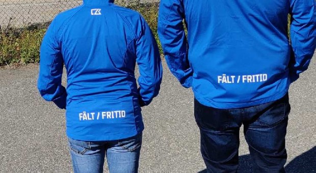 Bild på ryggtavlan på två personer som bär blåa jackor med texten Fritid/Fält